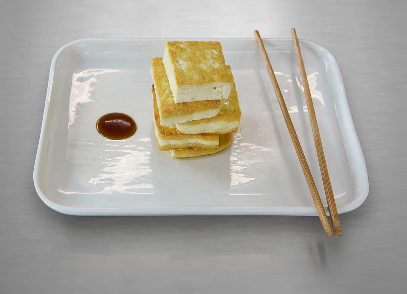 Gestapelter gebratener Tofu mit Soße und Essstäbchen auf einem weißen Teller.